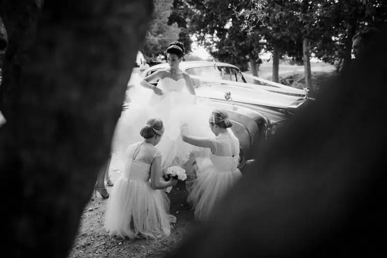 Black-and-white view of bridesmaids fluffing the bride's skirt beside the car