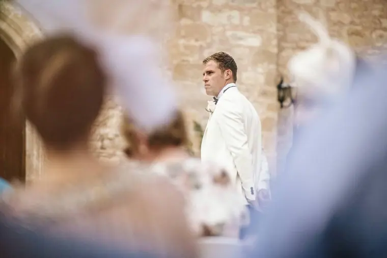 Groom watching his bride approach, seen past blurred guests' hats