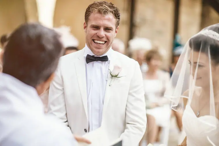 Groom in a white tuxedo jacket beaming at his bride during the ceremony