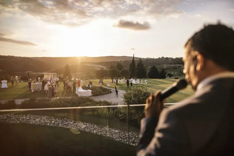 Man speaking into a microphone on the terrace as guests gather on the lawn at sunset