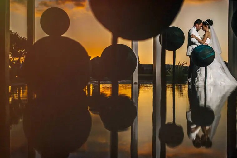 Bride and groom kissing behind giant balloons reflected in a dark pool at sunset
