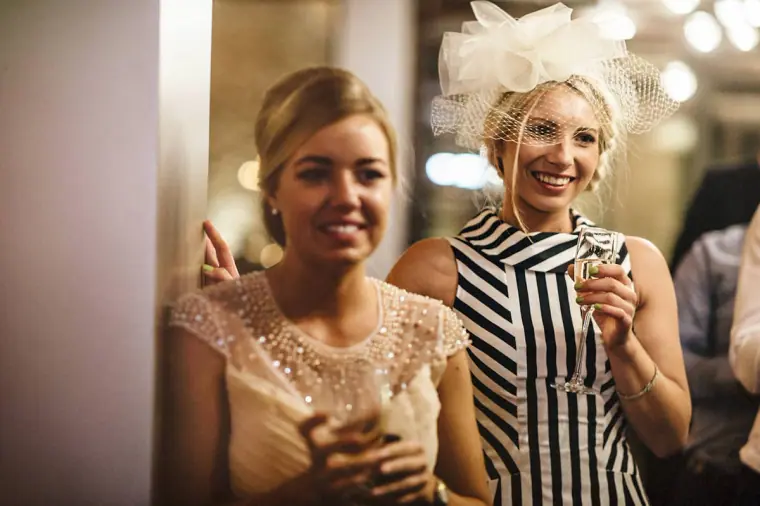 Two smiling female guests, one in a striped dress and fascinator, holding drinks