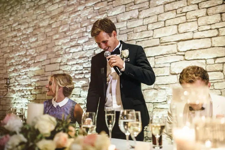 Groom giving his speech in front of a stone wall as guests listen at the top table