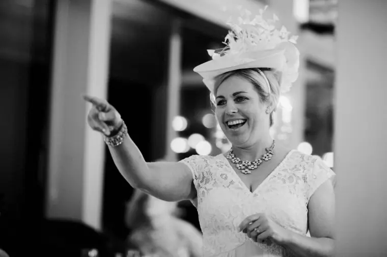 Black-and-white shot of a laughing guest in a fascinator pointing during the speeches