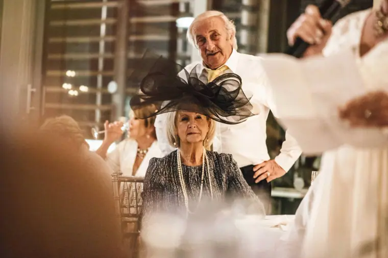 Older guest in a black fascinator smiling as a man reads a speech beside her