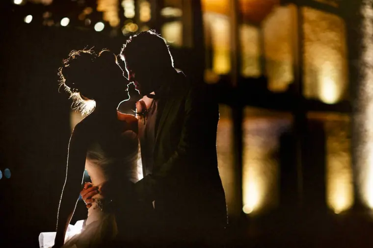Silhouetted bride and groom about to kiss against the warm glow of the venue at night