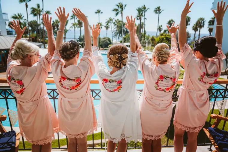Bride and bridesmaids in matching pink robes cheering with arms raised by the pool