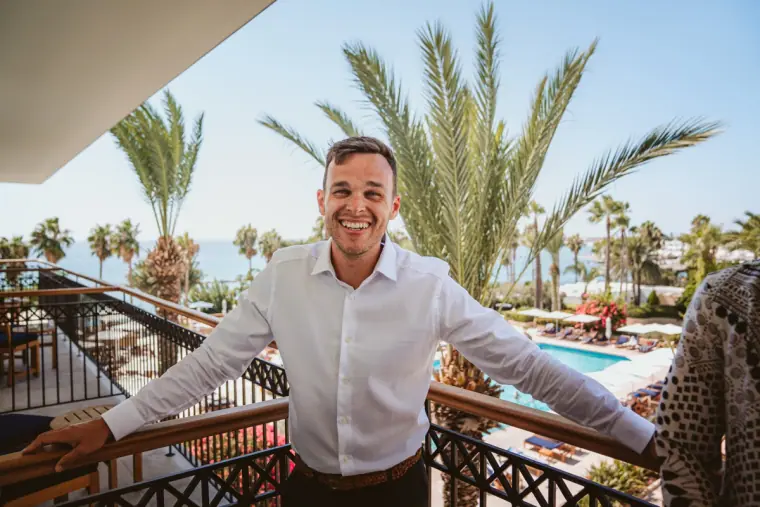 Groom grinning on a hotel balcony with palm trees and pool behind