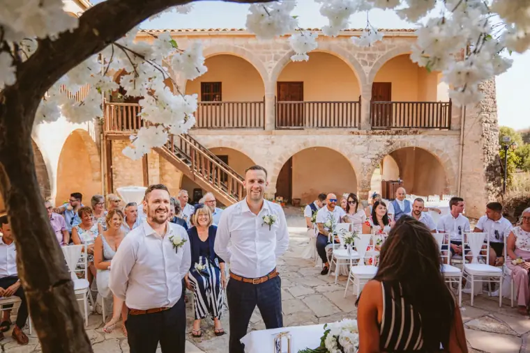Groom and best man waiting under blossom trees as guests take their seats