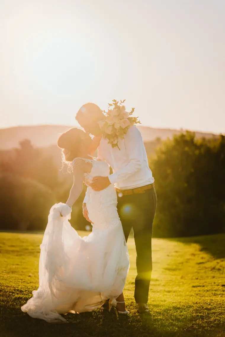 Bride and groom kissing on a sunlit lawn, her dress swept up in the breeze