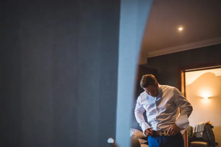 Groom buttoning his shirt in a warmly lit hotel room