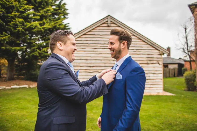 Best man adjusting the groom's buttonhole on the lawn by a wooden barn
