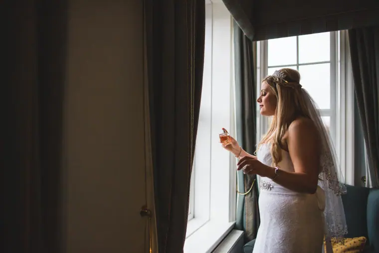 Bride putting on her earring by a tall window in the hotel room