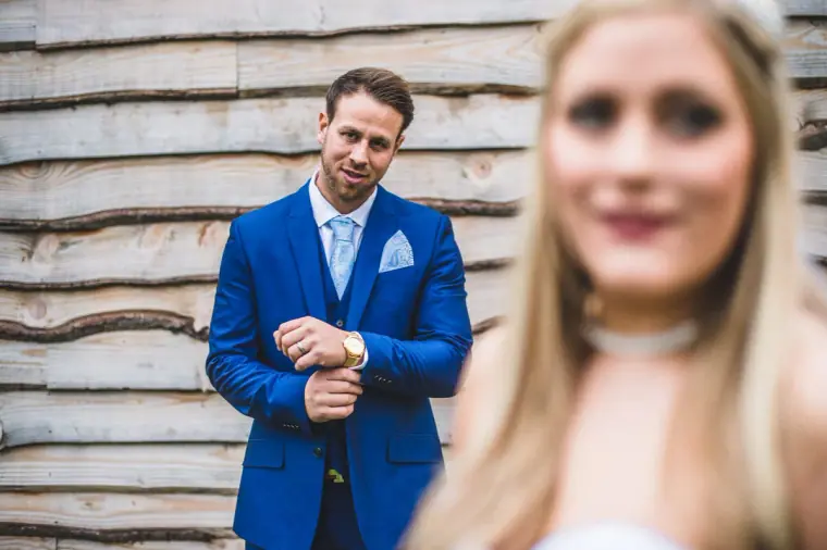 Groom in a bright blue suit by a timber wall, the bride blurred in the foreground