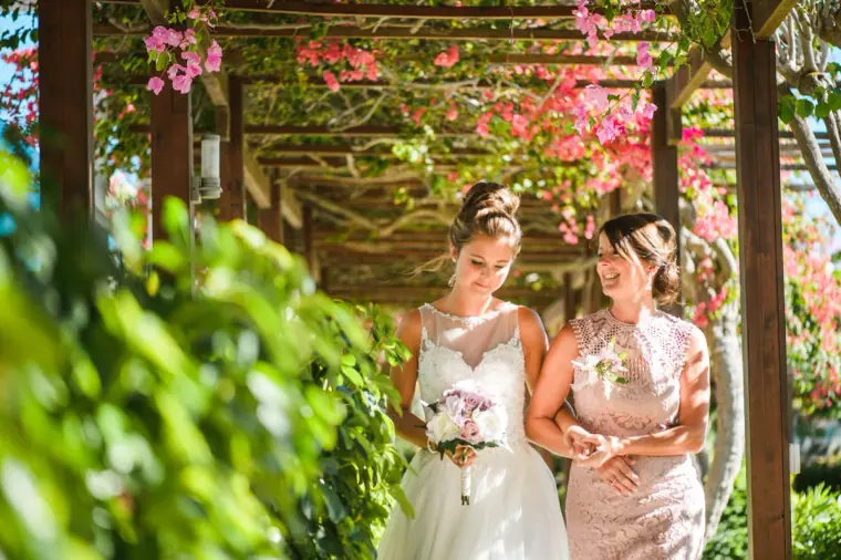 Bride walking beneath the bougainvillea pergola with a woman in blush lace