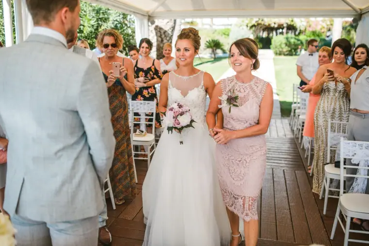 Bride walked down the aisle by a woman in blush lace as the groom waits