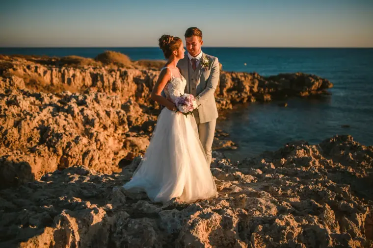 Bride and groom standing on rocky coastline by the sea at sunset, Ayia Napa