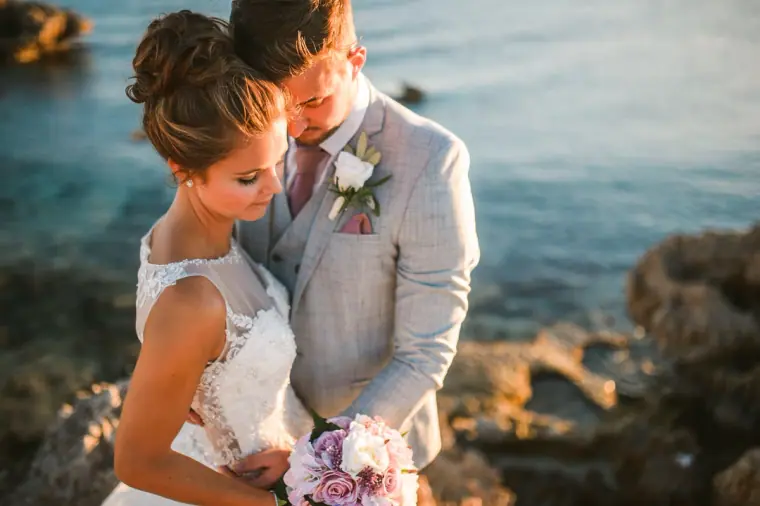 Groom kissing his bride's head on the rocks by the sea at golden hour