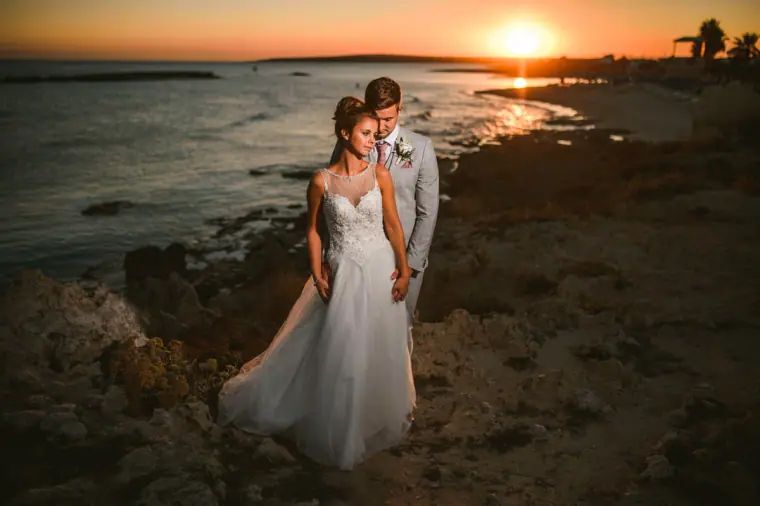 Bride and groom standing on the shoreline rocks at sunset
