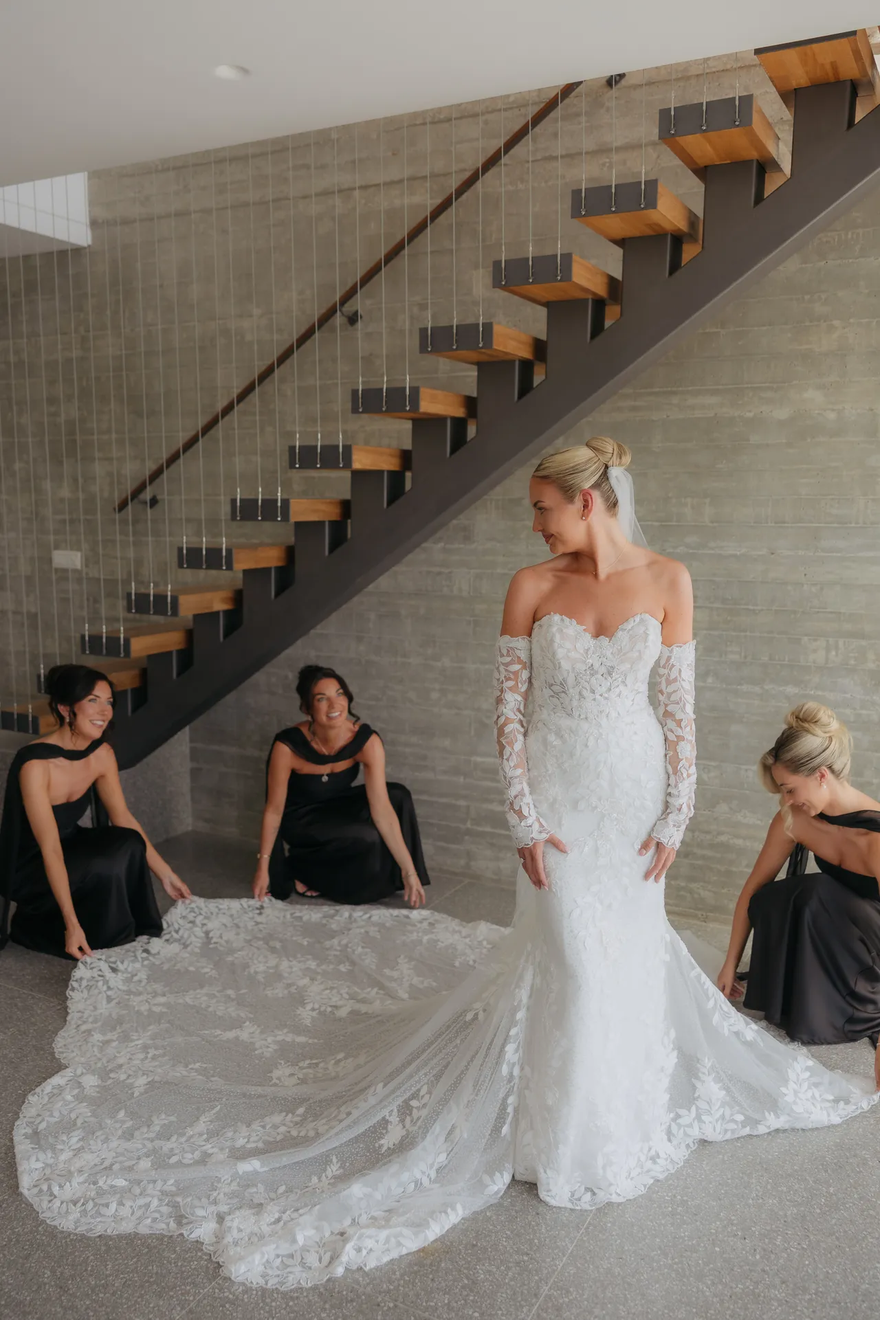 Bride in a lace mermaid gown with a long train as bridesmaids look on by the stairs