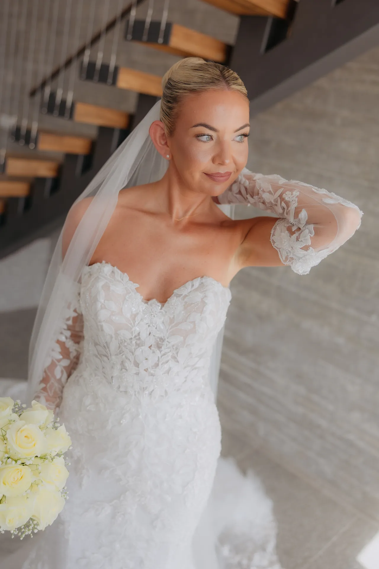 Olivia in her strapless lace gown and veil touching her earring by the staircase