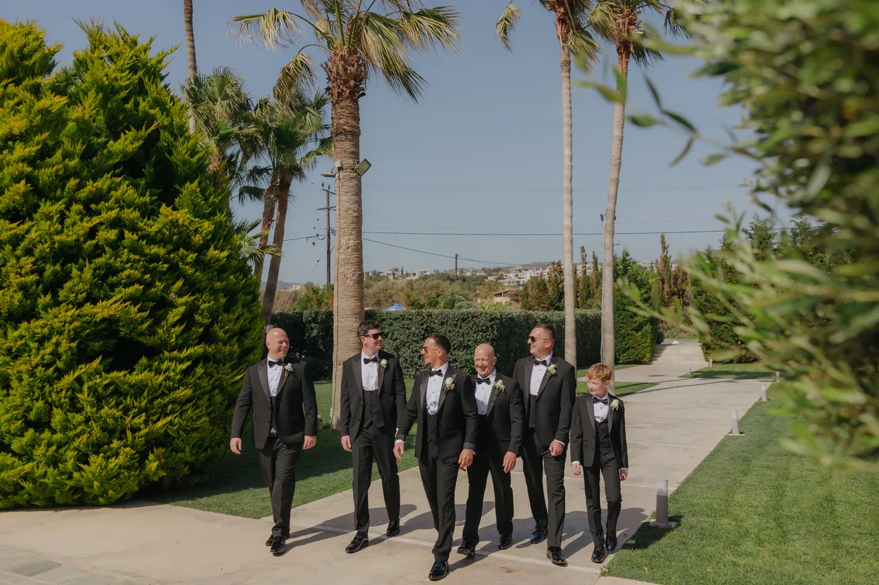 Groom and groomsmen in tuxedos walking together along the palm-lined path