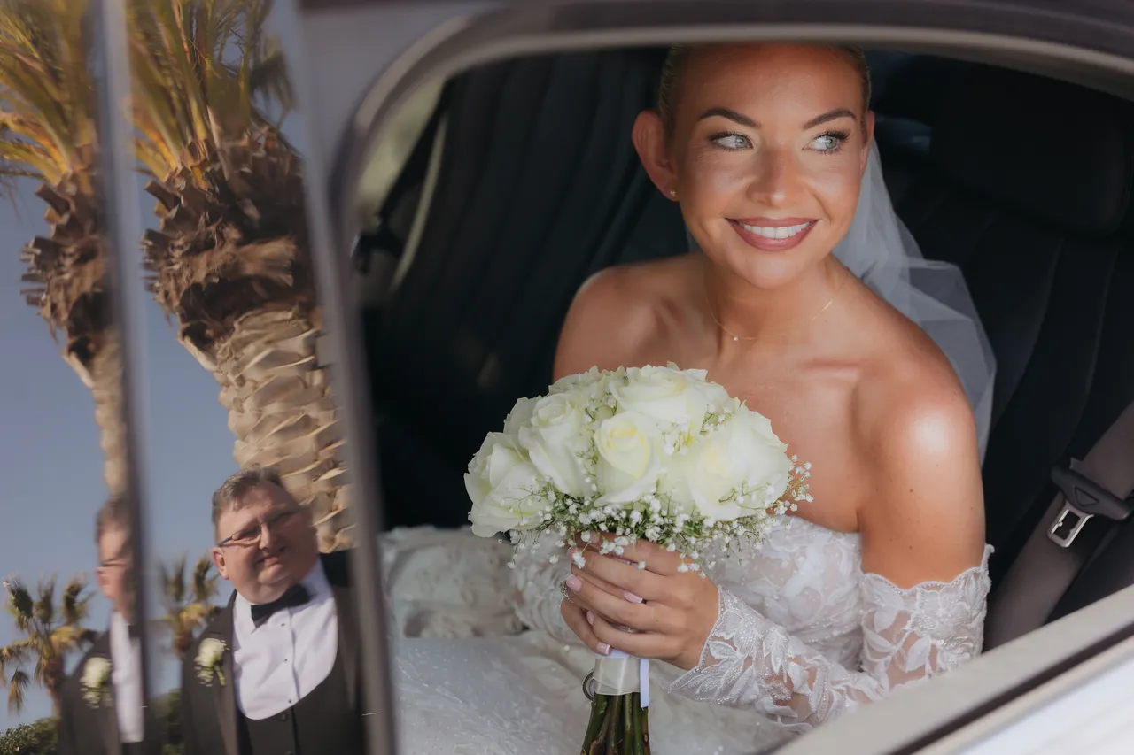 Olivia smiling from the car with her white rose bouquet as she arrives