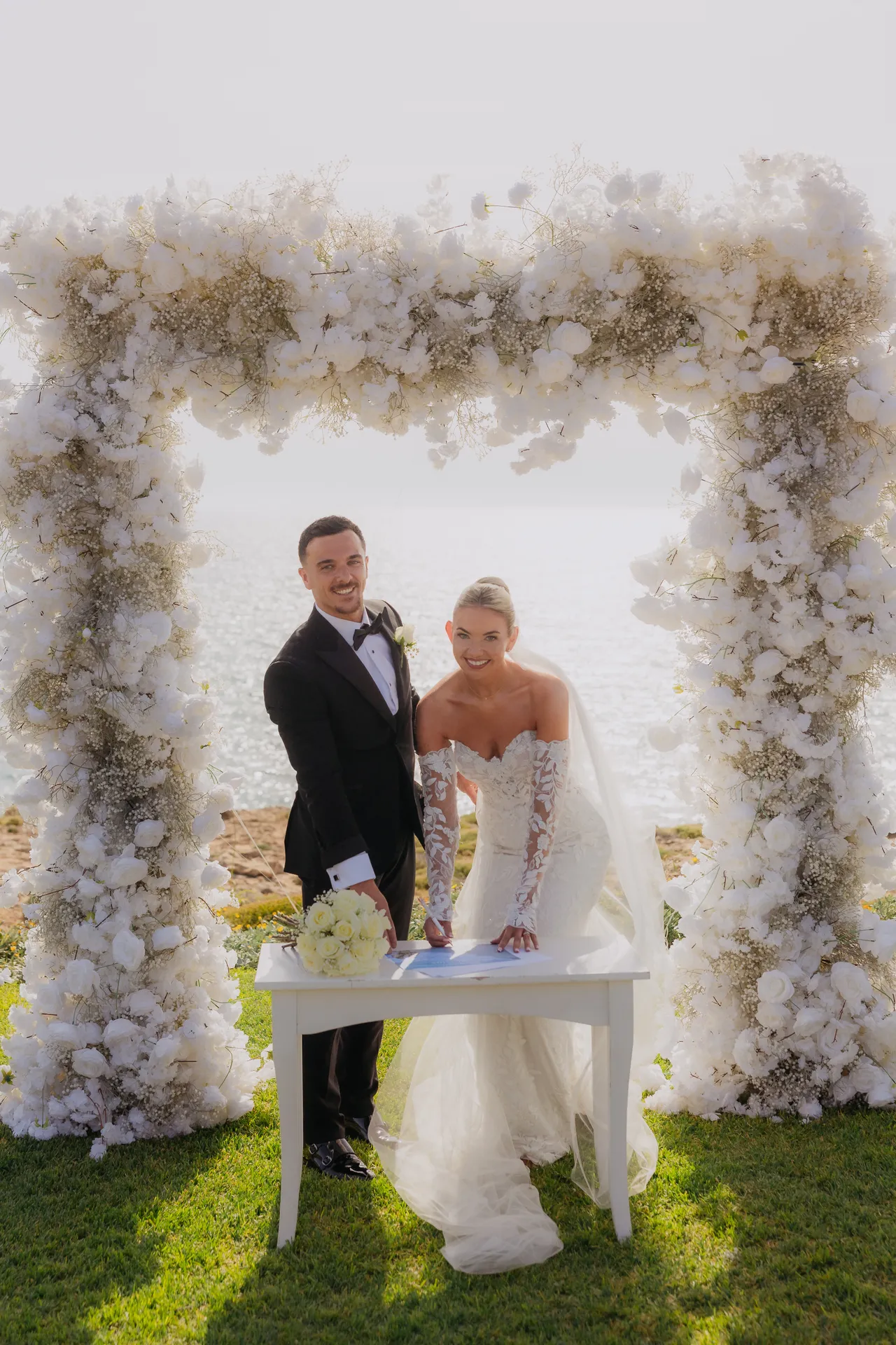 Olivia and Ashley signing the register under a white floral arch by the sea