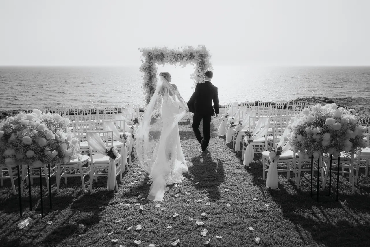 Black-and-white photo of the couple walking towards the floral arch above the sea