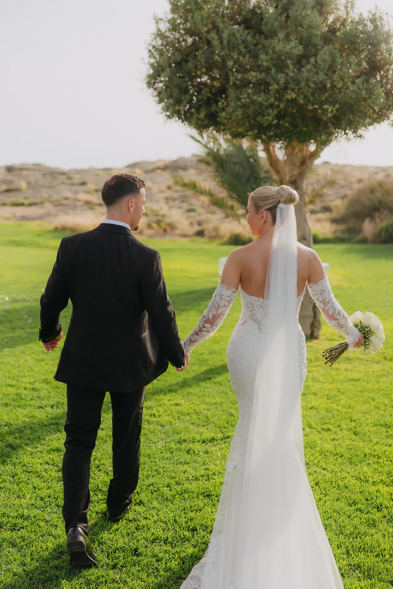 Bride and groom walking hand in hand across the lawn towards an olive tree