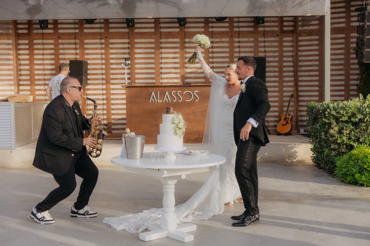 Saxophonist dancing beside the couple and their cake in front of the Alassos sign