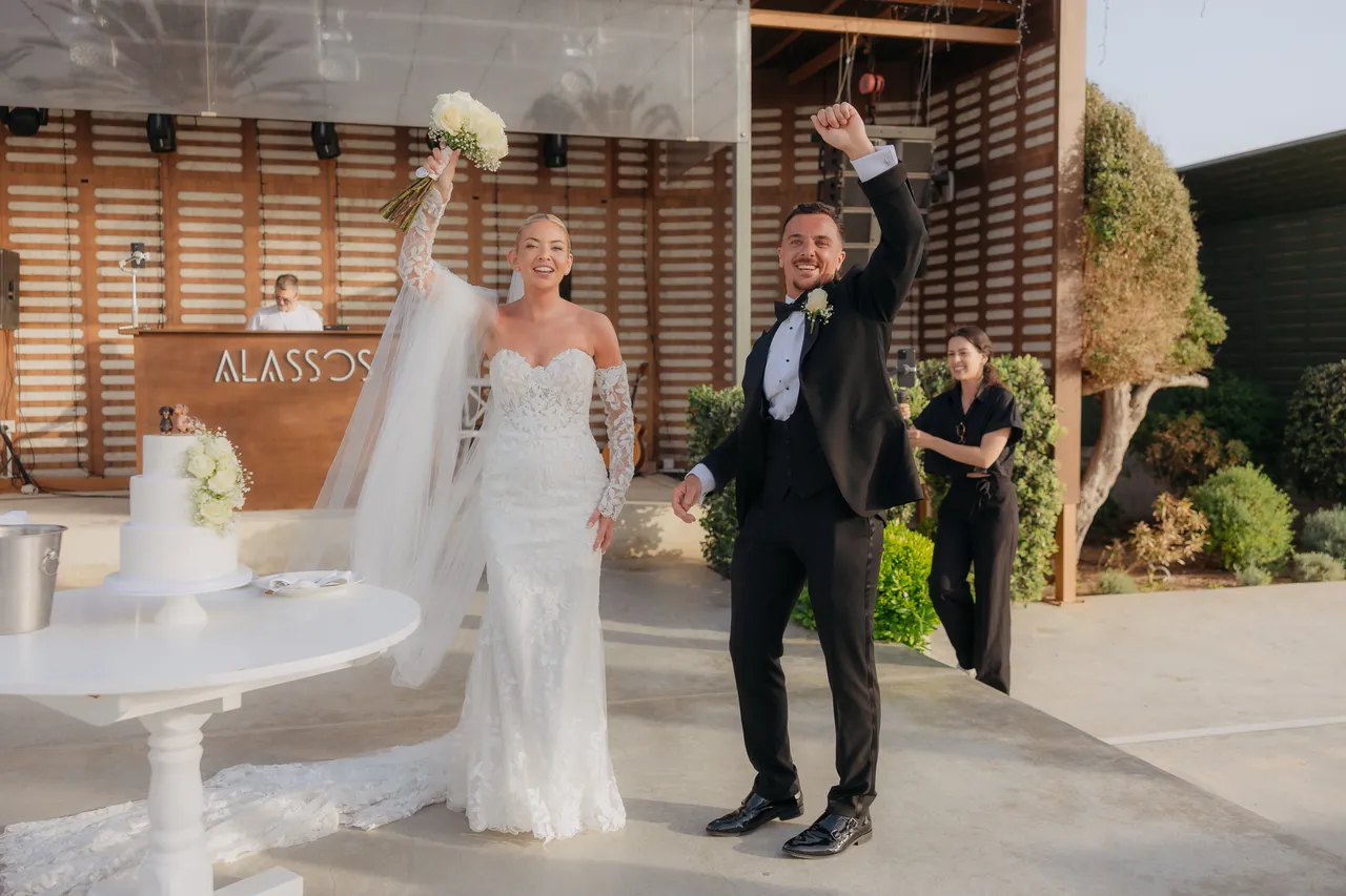 Olivia and Ashley celebrating with arms raised beside their wedding cake at Alassos