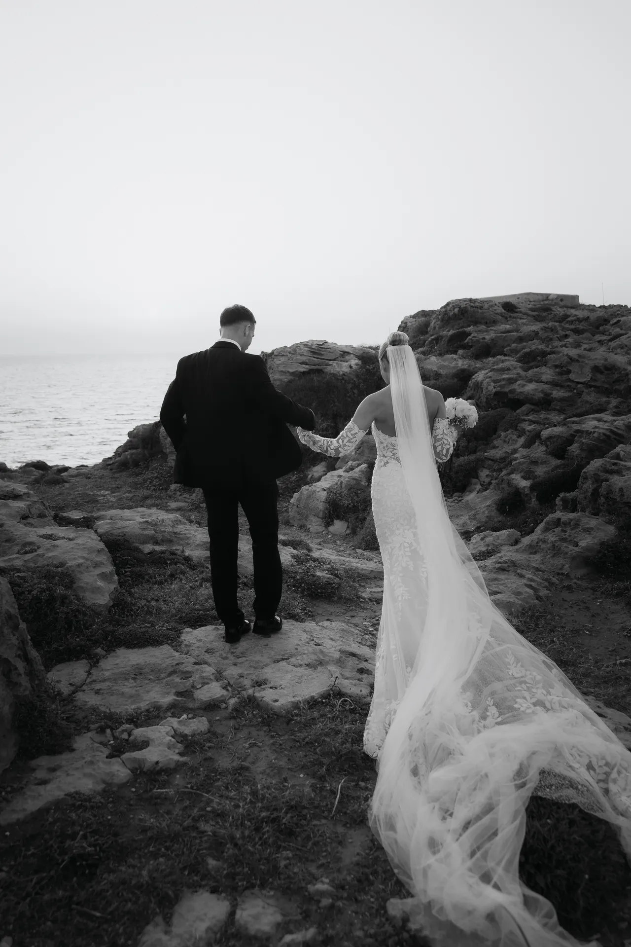 Black-and-white photo of the couple walking over rocks, veil trailing behind