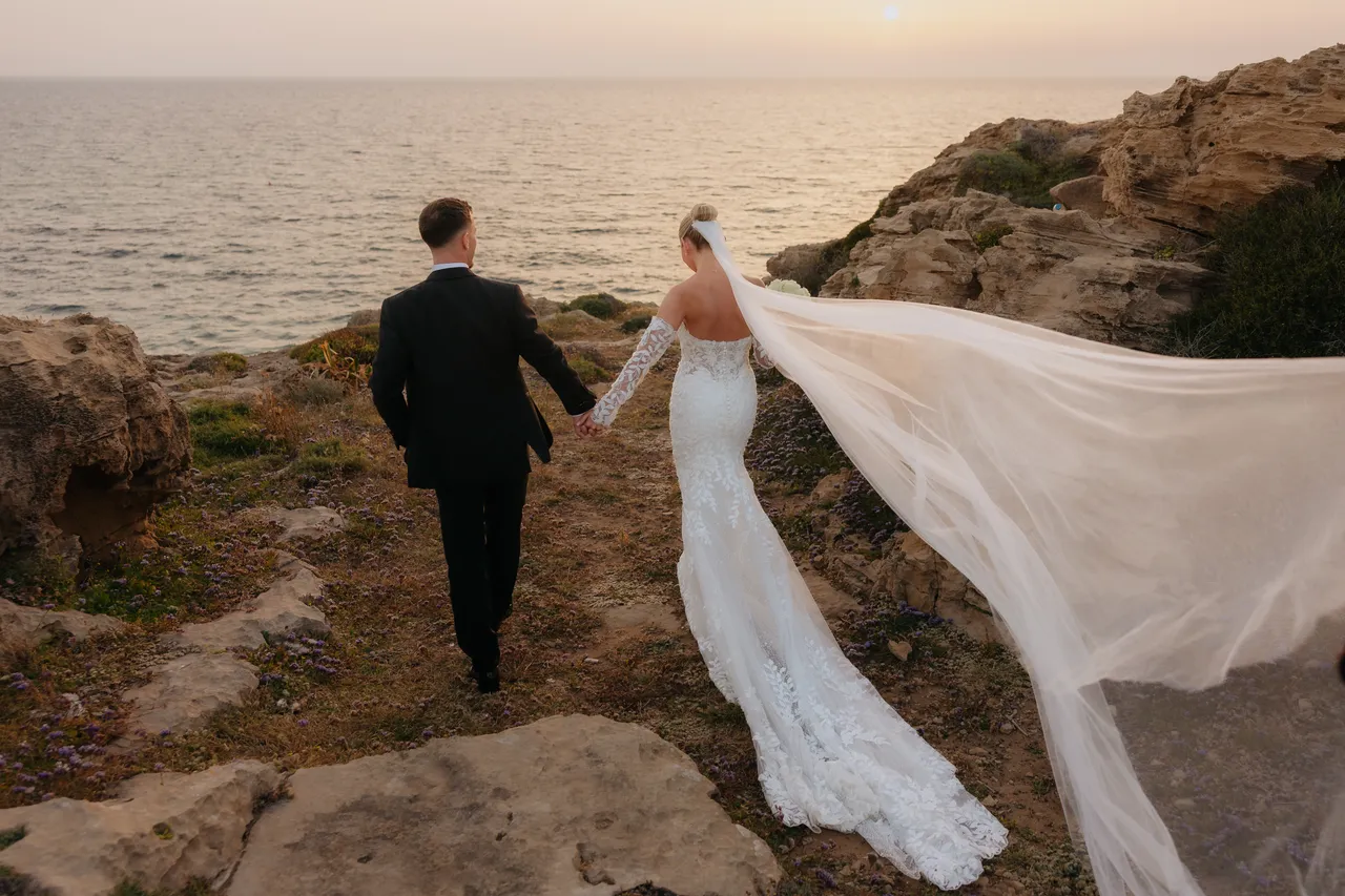 Couple walking hand in hand along the clifftop, veil streaming in the wind