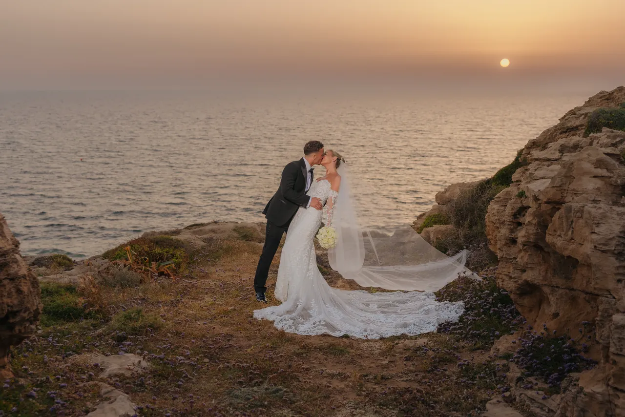 Bride and groom kissing on the rocky shore as the sun sets over the sea