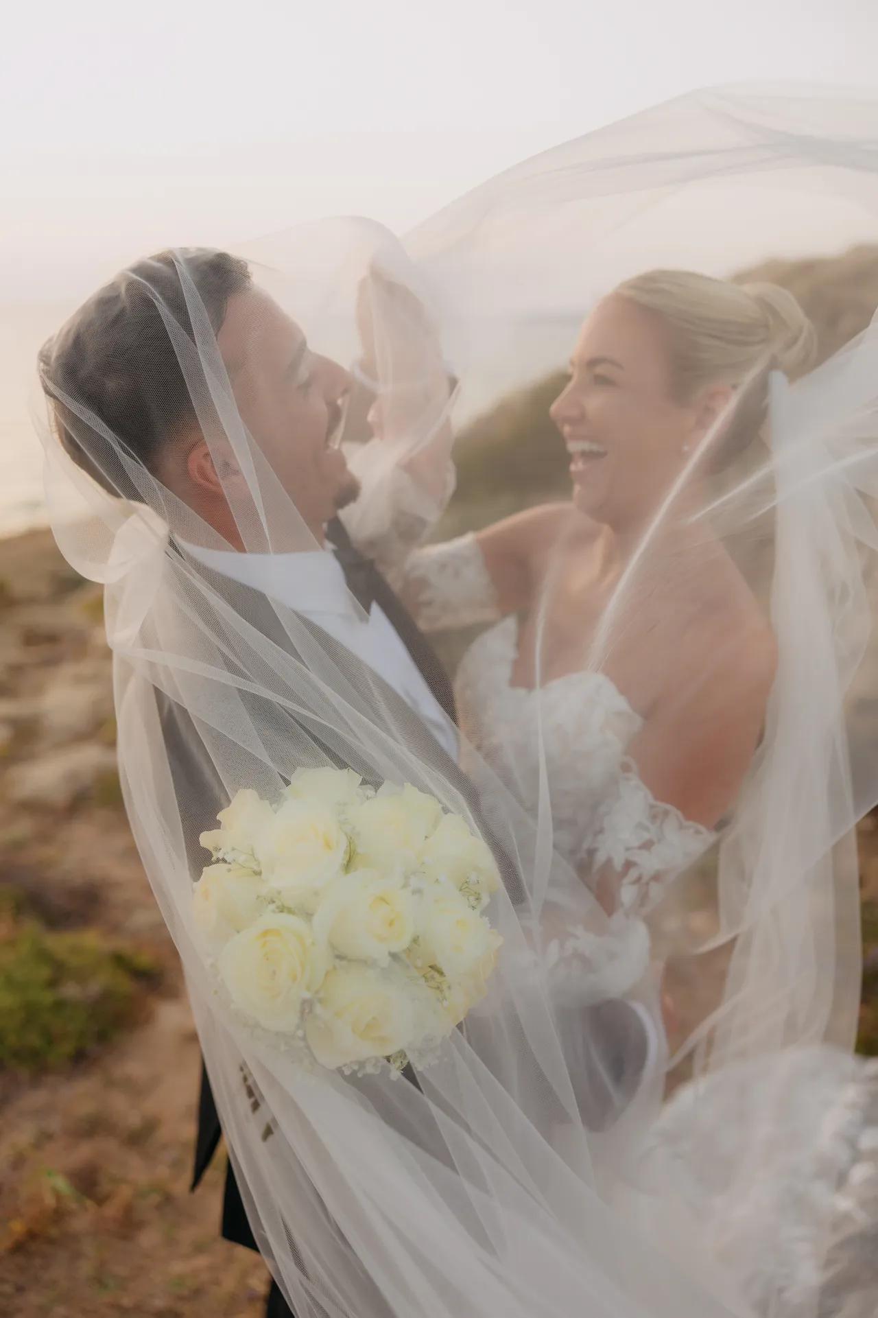 Couple laughing beneath the bride's veil, her white rose bouquet in hand