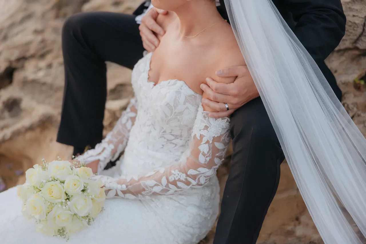 Groom's arms around his bride as she holds white roses on the rocks