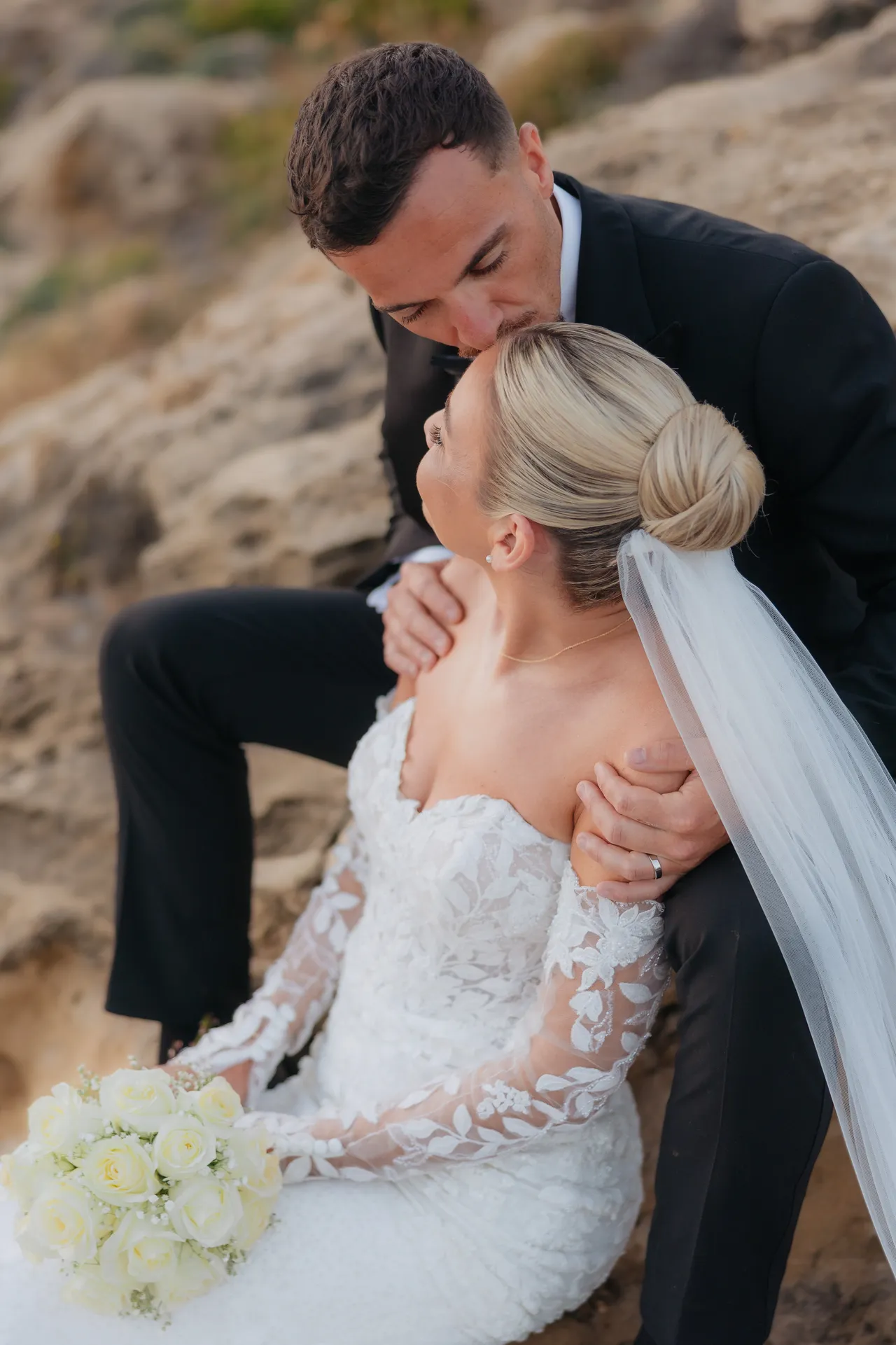 Groom kissing the top of his bride's head as they sit on the rocks