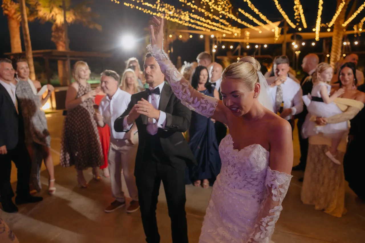 Bride dancing with her arm raised among guests under string lights