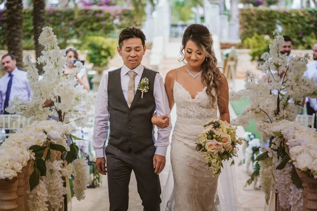 Bride walking down an aisle lined with white orchids, escorted by a man in a grey waistcoat