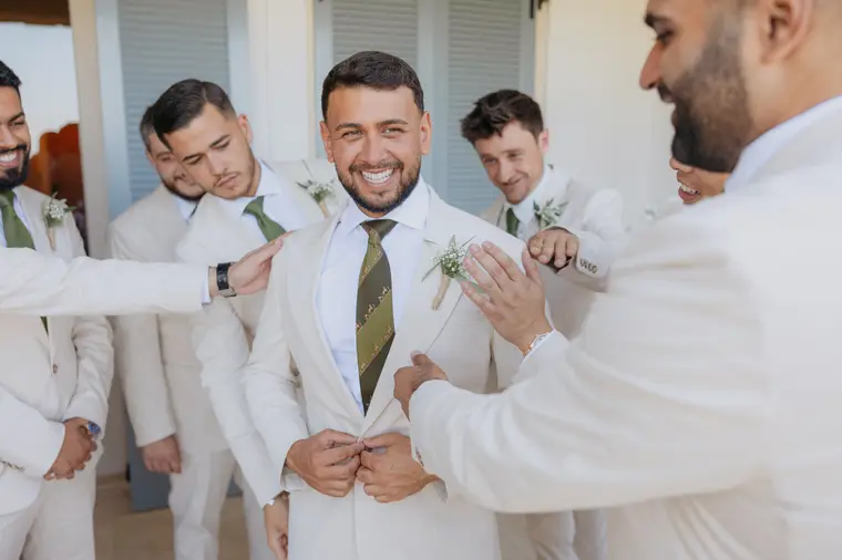 Groomsmen buttoning Andrew's cream jacket as he laughs
