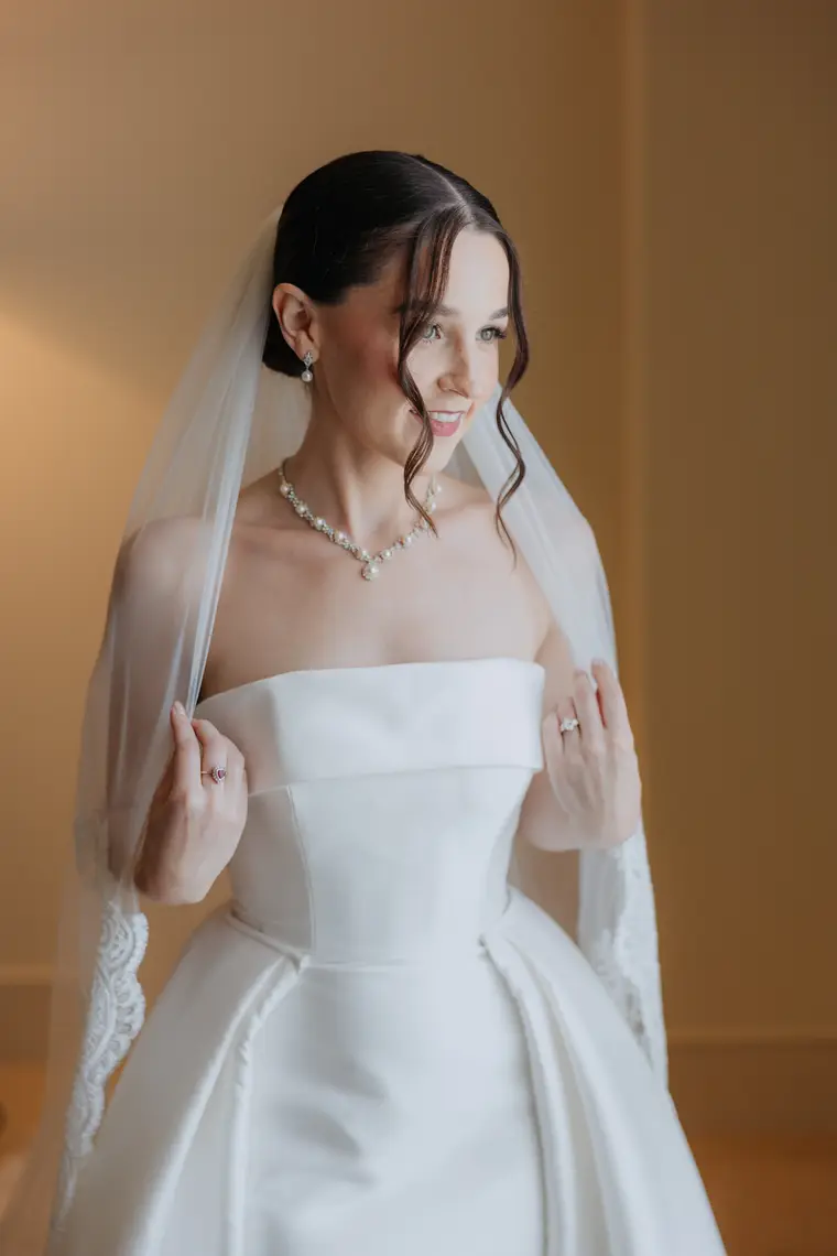Poppy in a strapless satin gown, veil and pearl necklace, getting ready