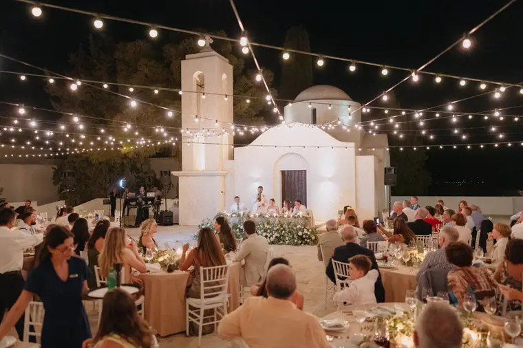 Guests dining beneath festoon lights in front of Anassa's white chapel