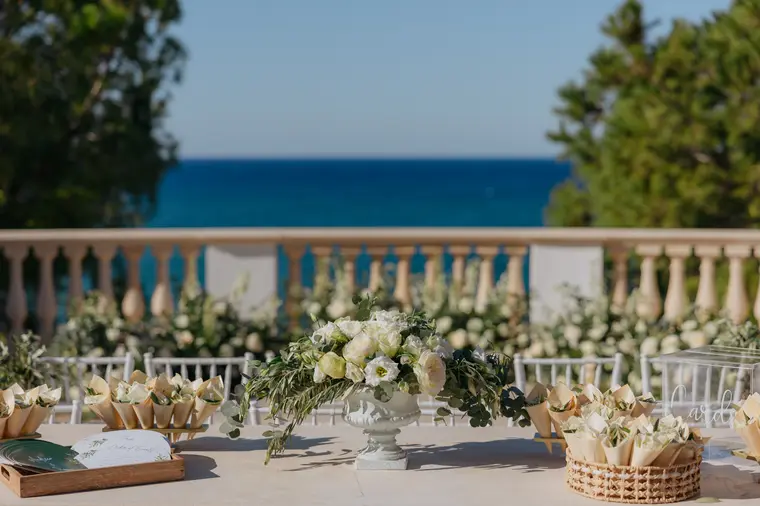 Ceremony table dressed with white florals and petal cones, the sea beyond the balustrade