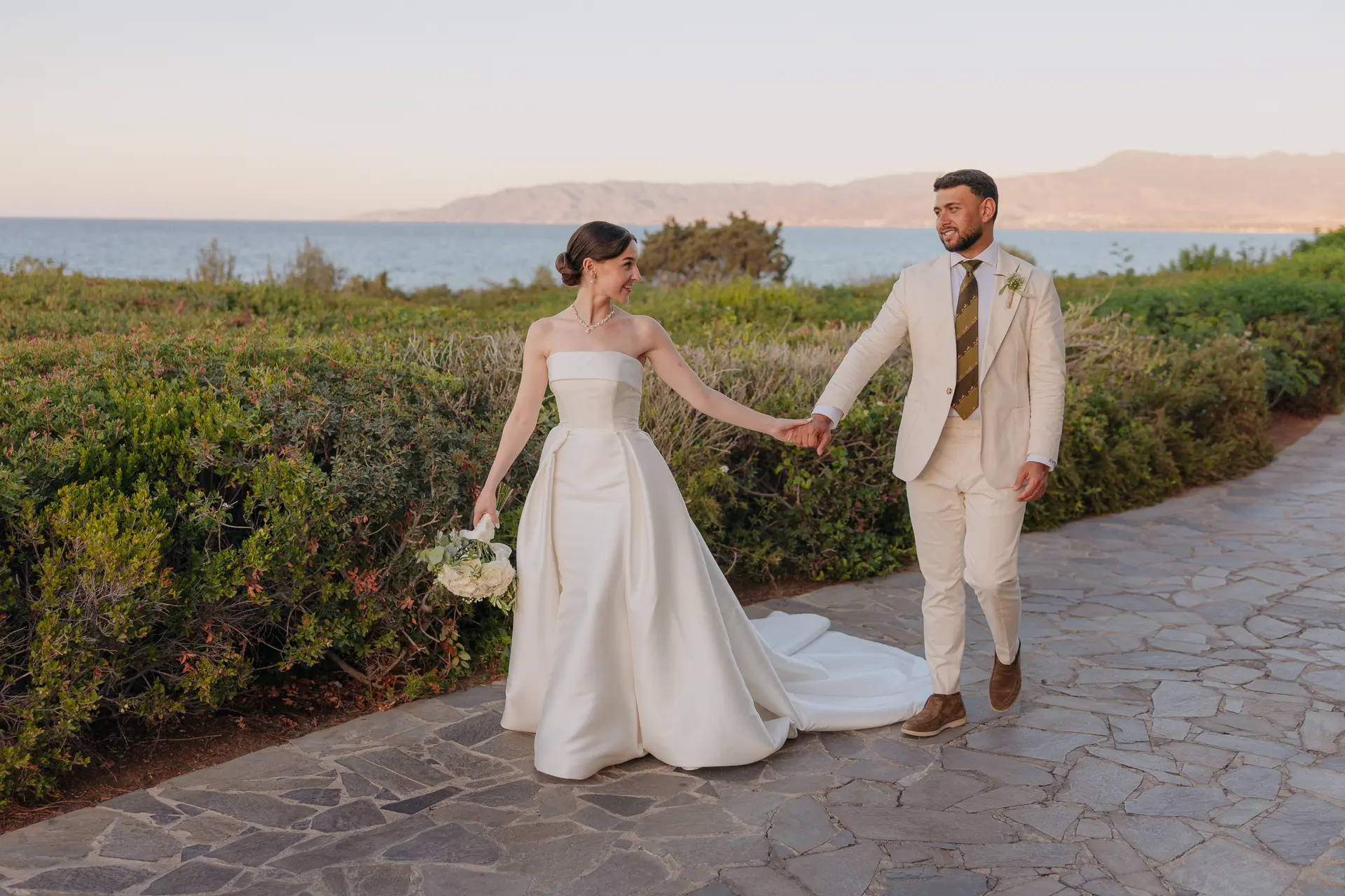 Couple walking hand in hand along a garden path, sea and mountains behind
