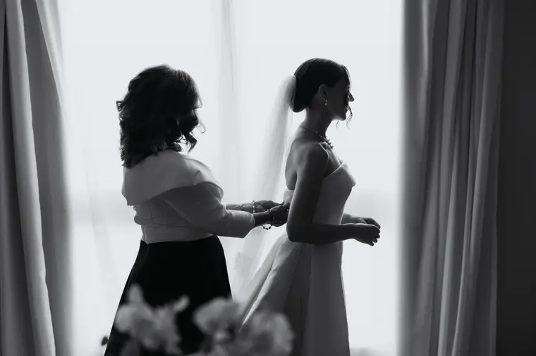 Black-and-white silhouette of a woman fastening the bride's dress by the window