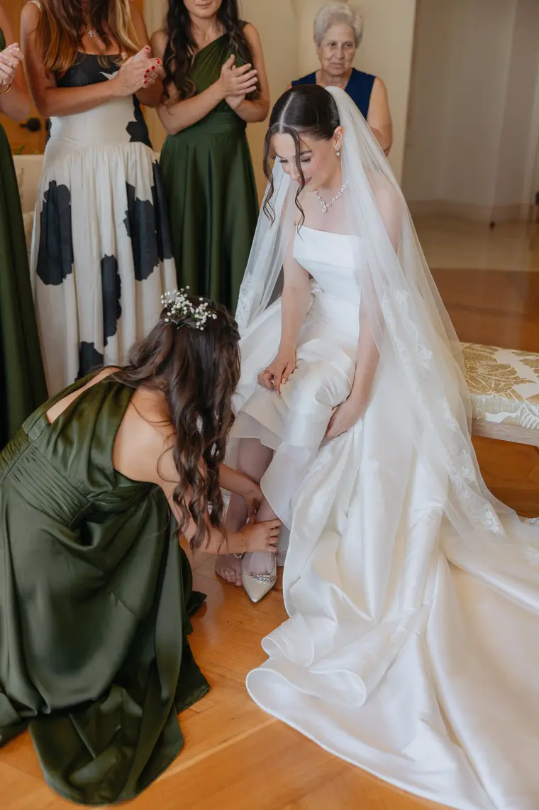 Bridesmaid fastening the bride's shoe as family looks on