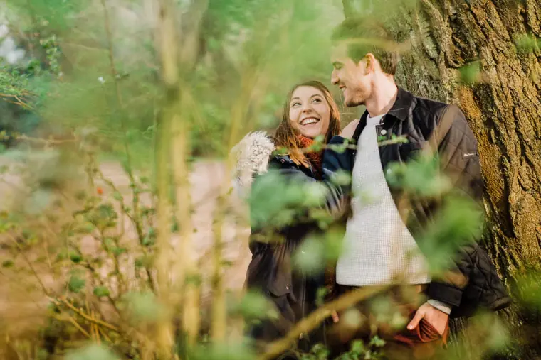 Couple laughing against a tree trunk framed by green foliage
