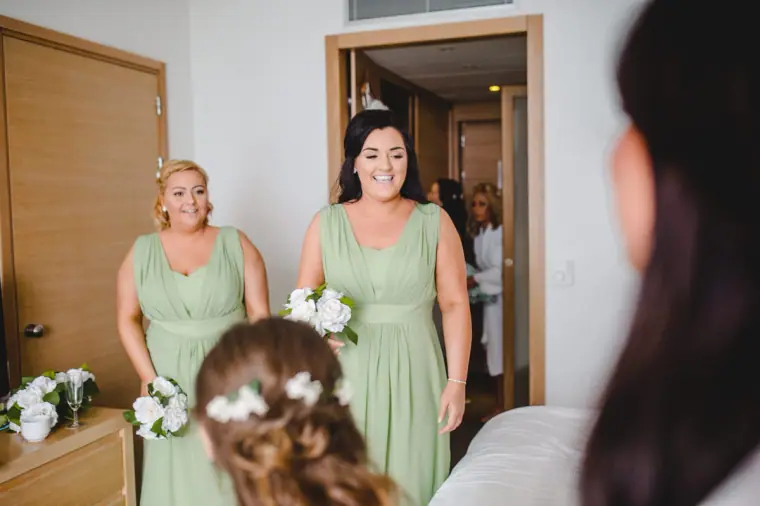 Bridesmaids in sage green holding white bouquets as the bride has her hair finished