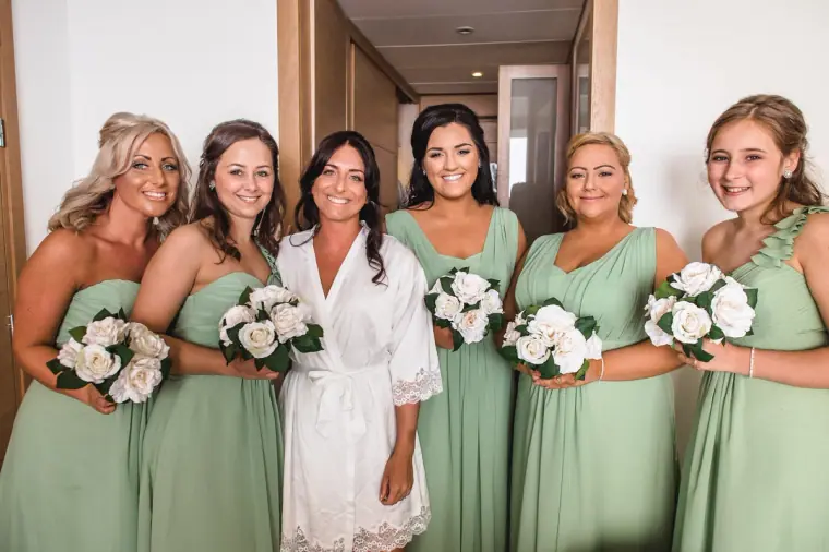 Bride in a lace robe with five bridesmaids in sage green holding white rose bouquets